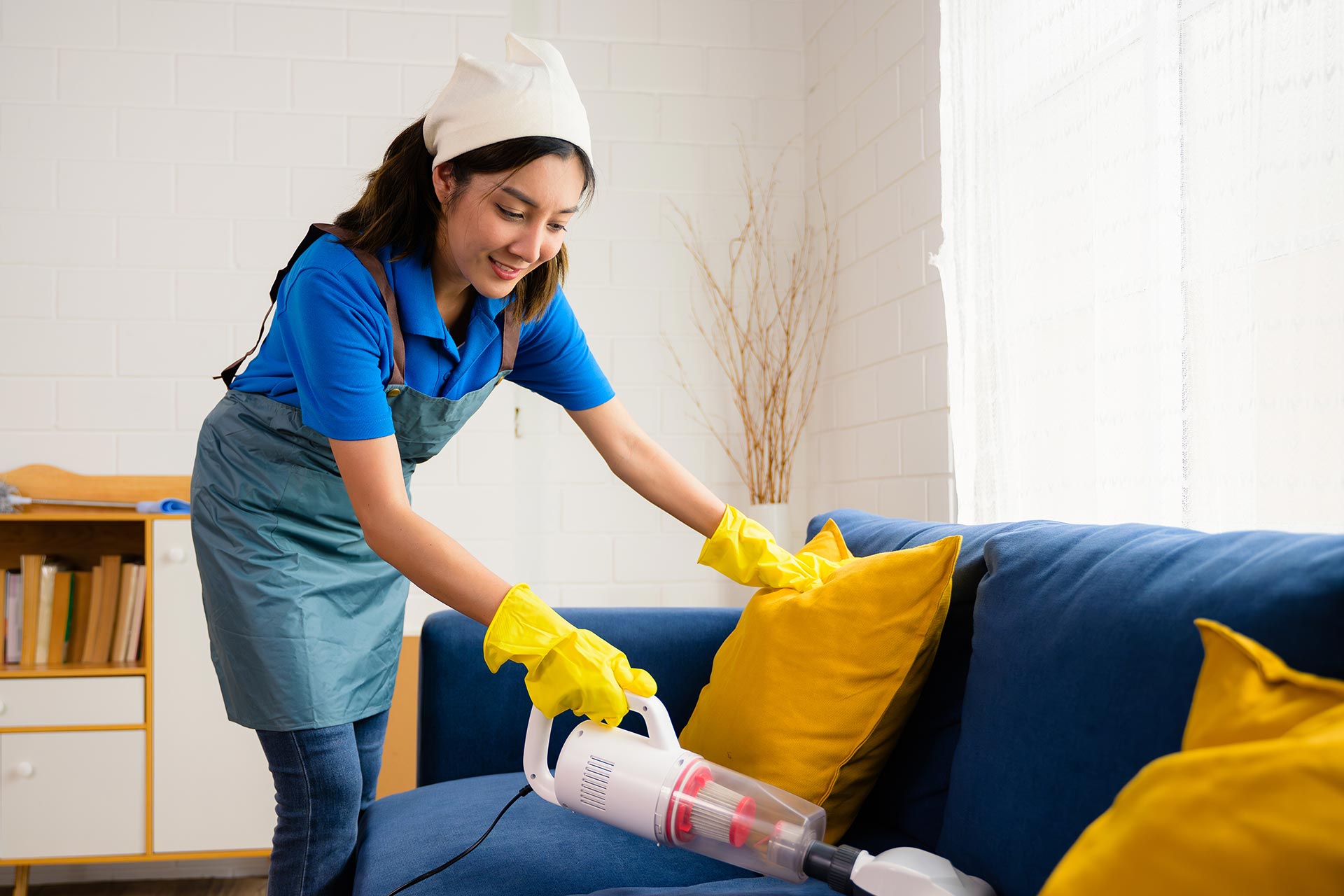 A woman wearing a blue shirt and apron is diligently cleaning a couch in a well-lit living room.